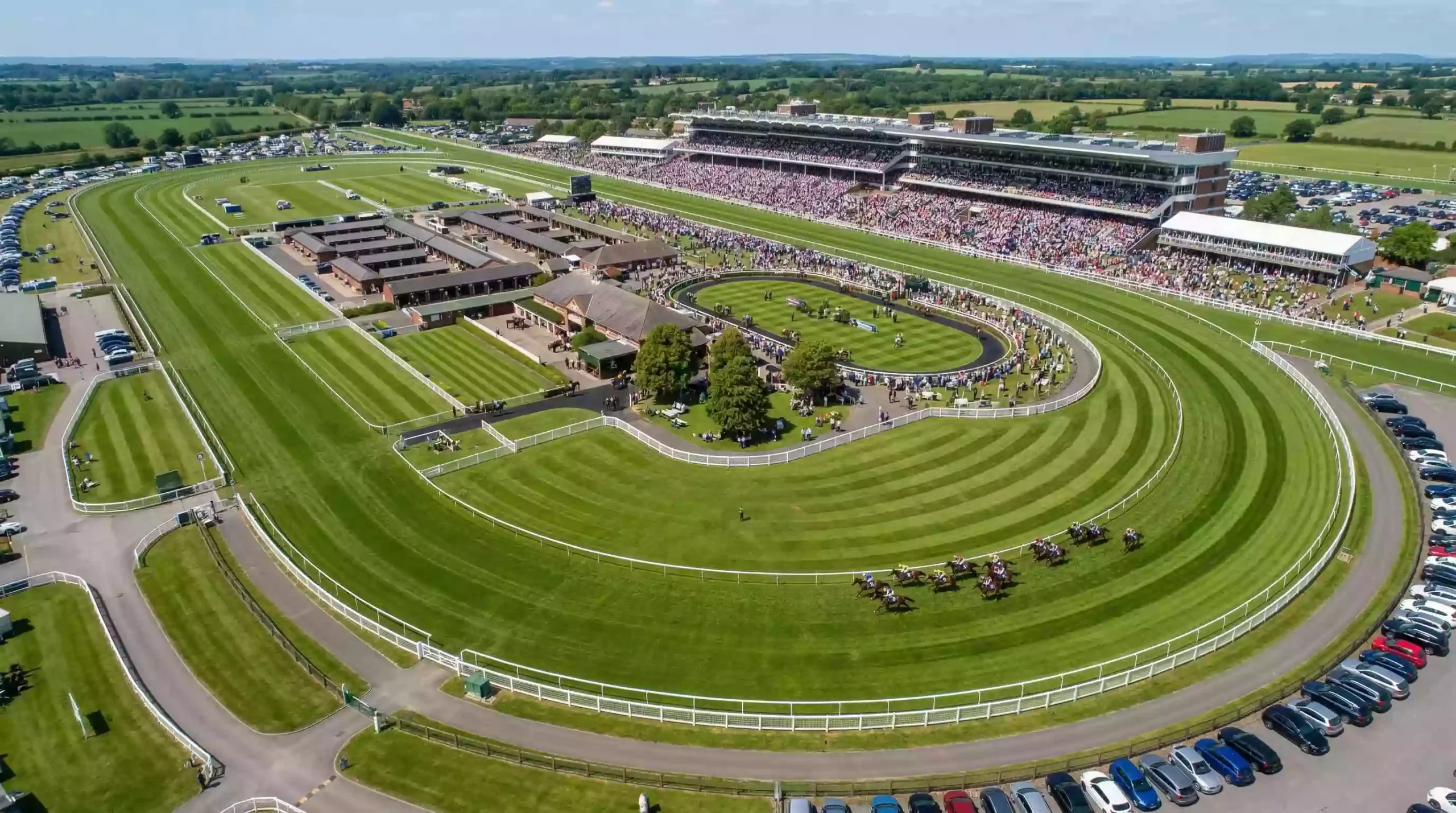 Aerial view of a UK racecourse with horses racing and a packed grandstand